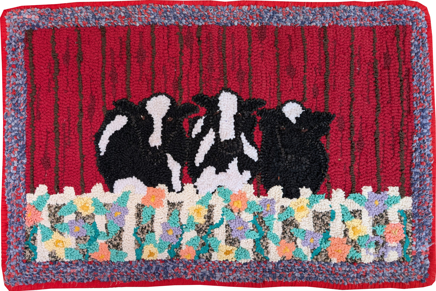 “Cow Klatch” – whimsical hand-hooked rug by Carmen Hall featuring black-and-white cows behind a floral fence on a red barn backdrop, outlined in dark green upholstery fabric.